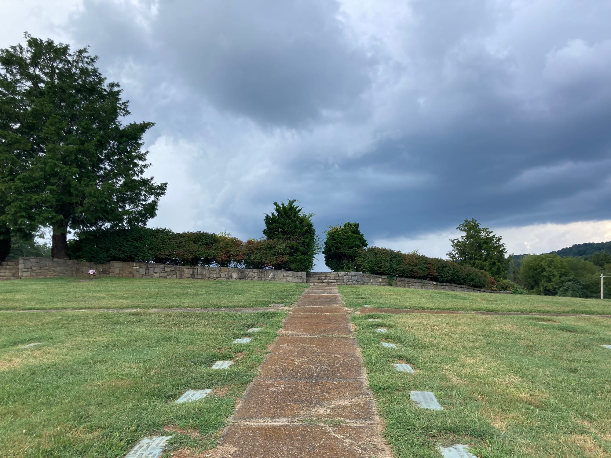 Sidewalk flanked by grave markers on grass leading to a stone fence and grove of bushes. 