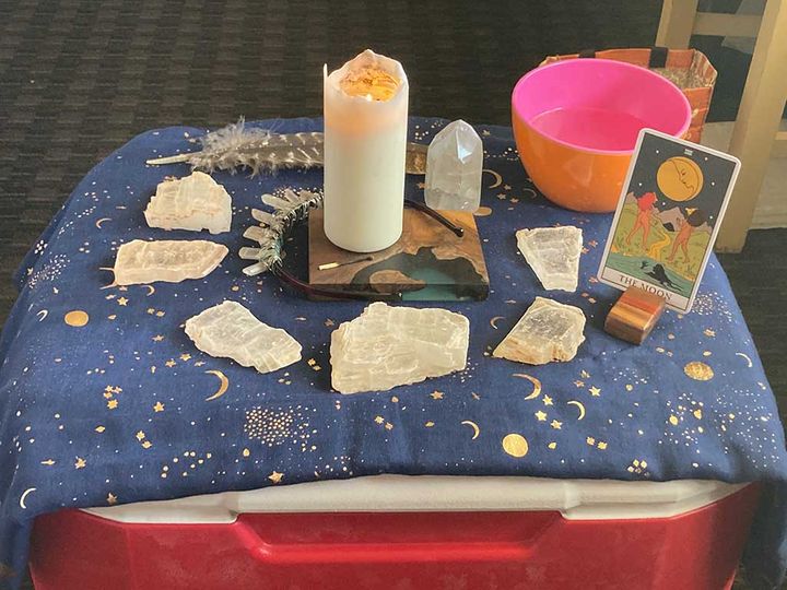 An altar with blue cloth, gold moon and stars, and crystals/candles/tarot cards set atop a red and white cooler. 