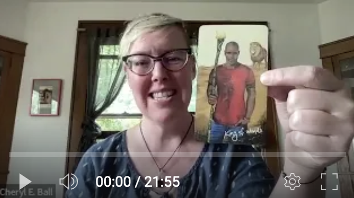 White woman with short blonde hair and glasses smiles and holds up a tarot card, the King of Wands