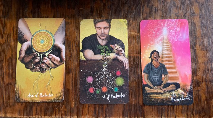 Three tarot cards -- Ace of Pentacles, 7 of Pentacles, and the Hierophant -- lay on a wooden table.