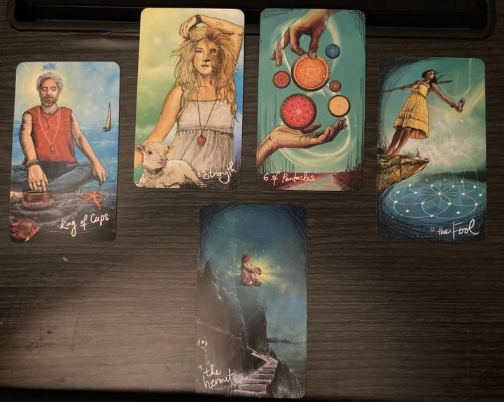 Five tarot cards spread out on a fake wooden laptop desk: Hermit, King of Cups, Strength, 6 of Pentacles, and the Fool