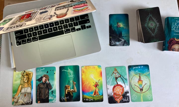 A laptop sits partially closed and surrounded by 7 tarot cards, a tarot deck, and the tarot guidebook.