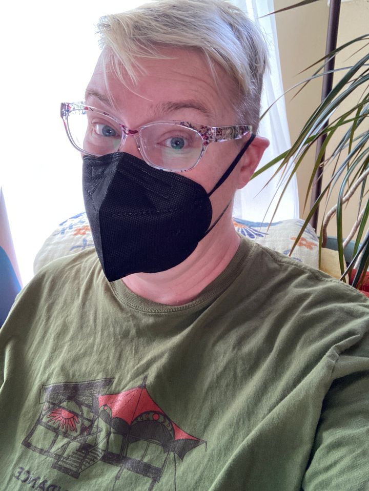 White woman with short blonde hair and glasses wearing a black mask and a green tee-shirt.