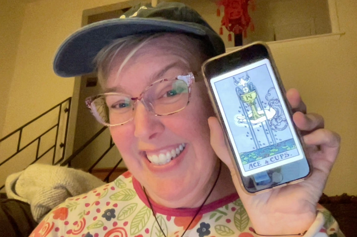 White blonde woman with blue baseball cap and multi-colored shirt grins at the camera holding her phone