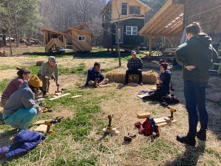 Seven people sit and stand in a circle on hay-strewn grass, with power drills and wood in front of them.